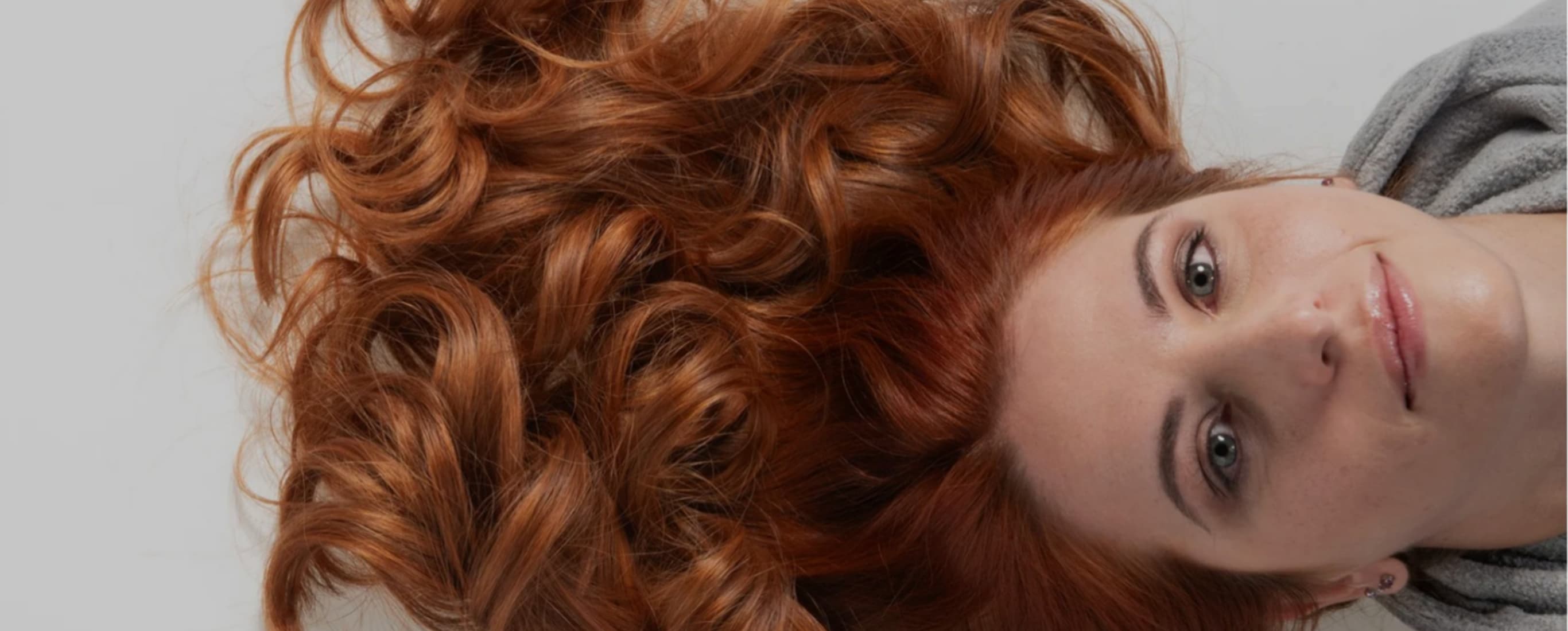 Donna sdraiata con lunghi capelli ricci ramati distesi in primo piano, espressione serena.