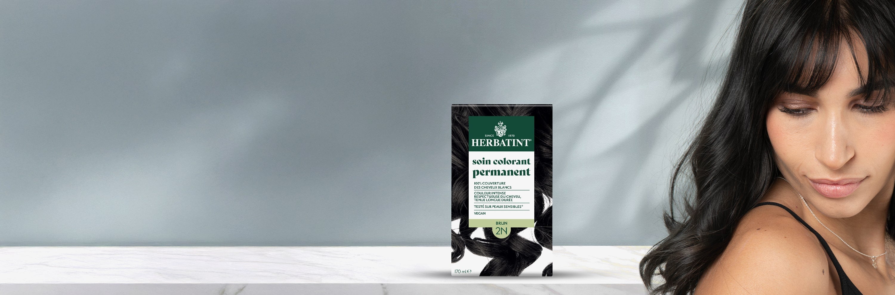 Boîte de gel colorant permanent Herbatint 2N châtain à côté d’une femme aux cheveux foncés, lisses et brillants.