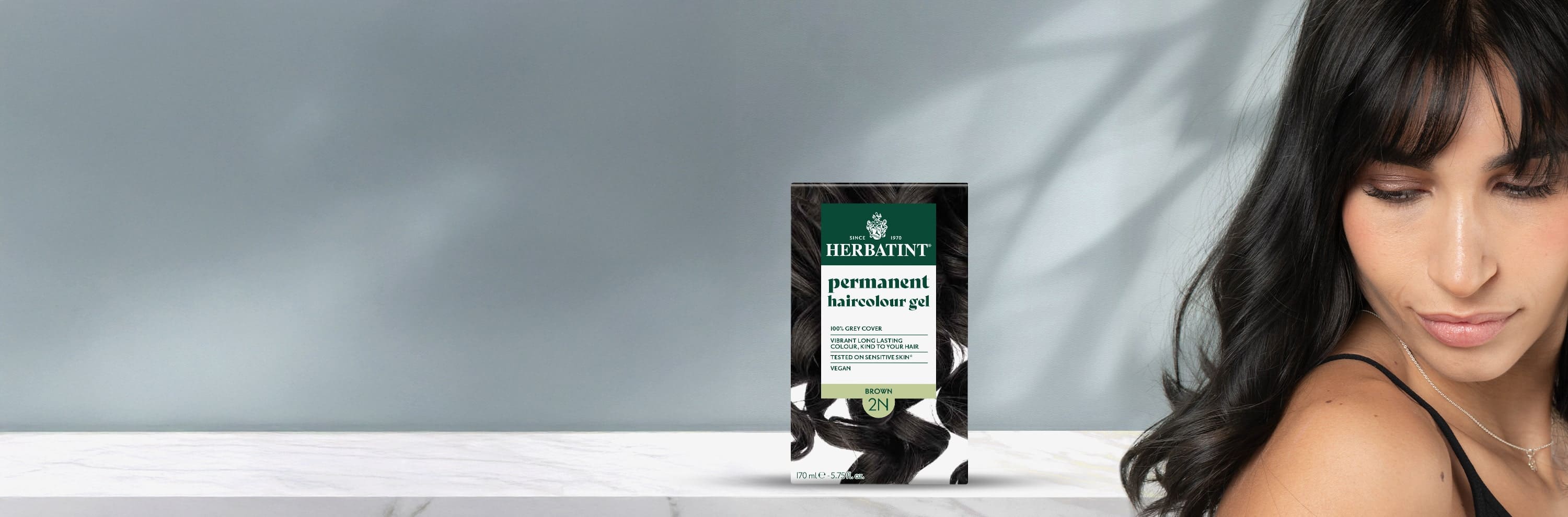 Verpakking van Herbatint 2N permanente kleurgel bruin naast een vrouw met steil, donker en glanzend haar.