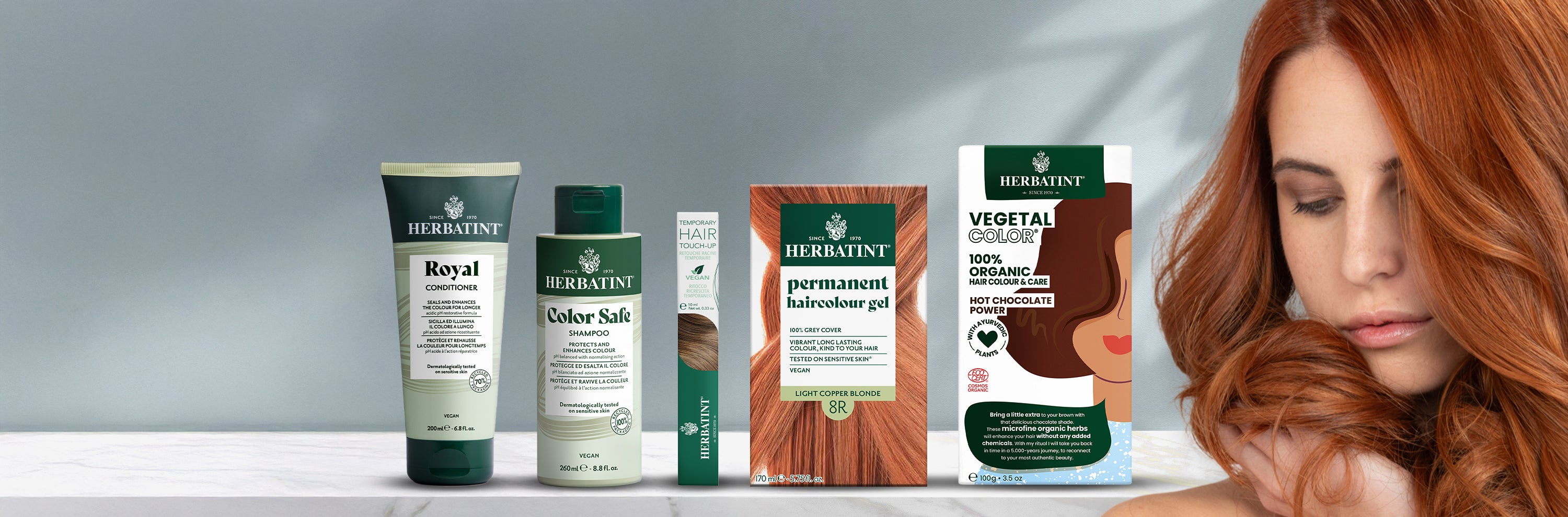 Volledige productlijn van Herbatint op een neutrale achtergrond getoond, inclusief shampoo, conditioner, haarkleurmascara, permanente kleuringsgels en Vegetal Color. Rechts streelt een vrouw haar lange koperkleurige haar.