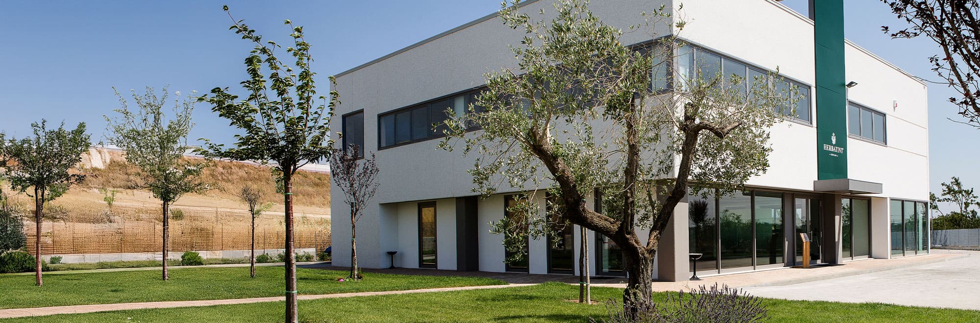 Vue extérieure du bâtiment principal de Herbatint, entouré d’arbres bien entretenus et d’espaces verts, avec le logo de l’entreprise sur un pilier vert vertical à l’entrée