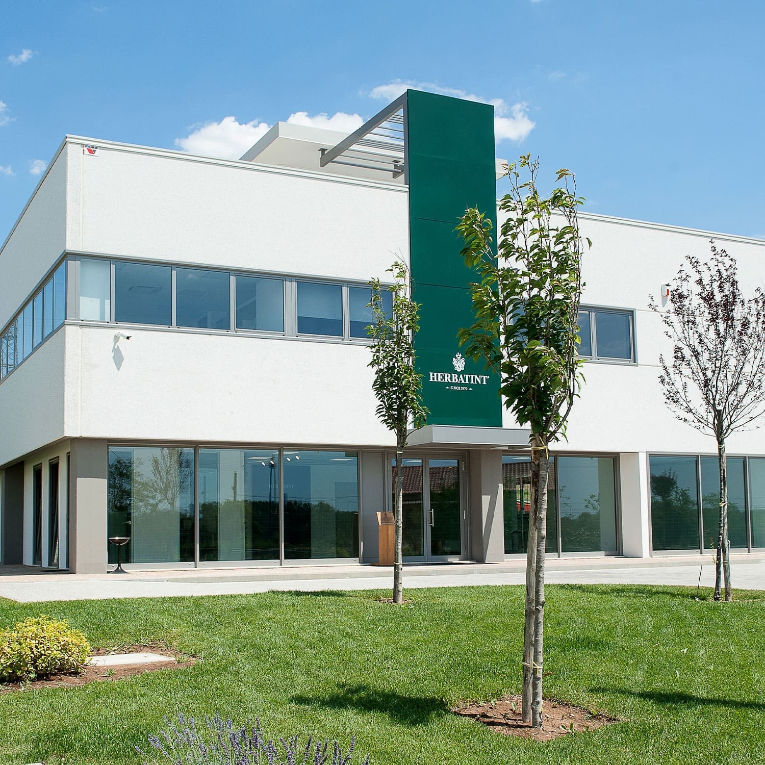 Entrada da sede da Herbatint com um jardim arborizado e o logótipo da empresa visível acima da porta