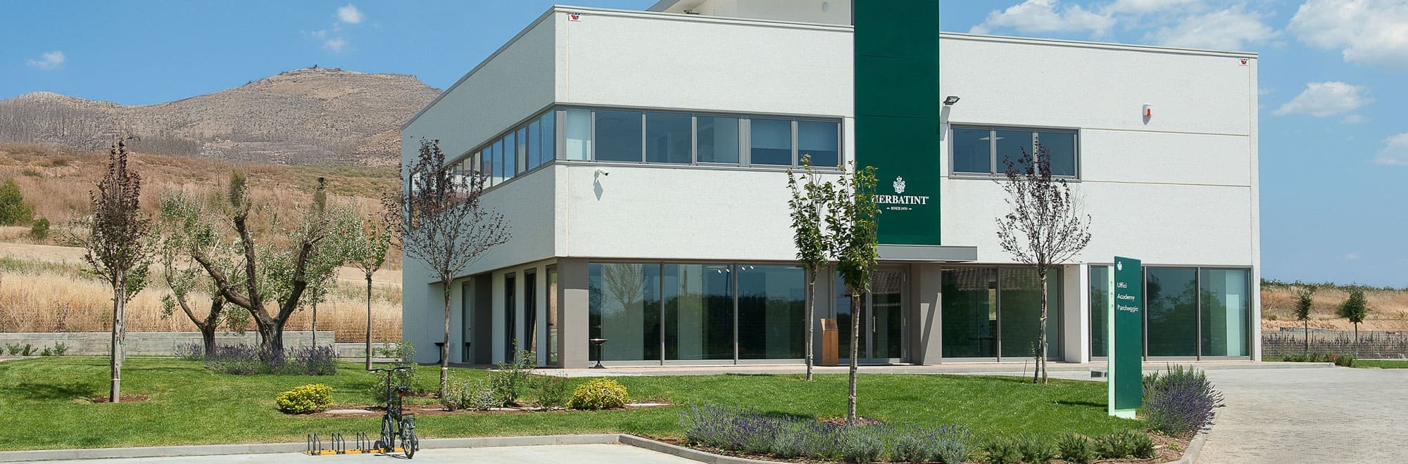 Vue extérieure du siège social de Herbatint, un bâtiment moderne de deux étages avec de grandes fenêtres et un logo vert vertical au centre de la façade. Le bâtiment est entouré d’un gazon bien entretenu