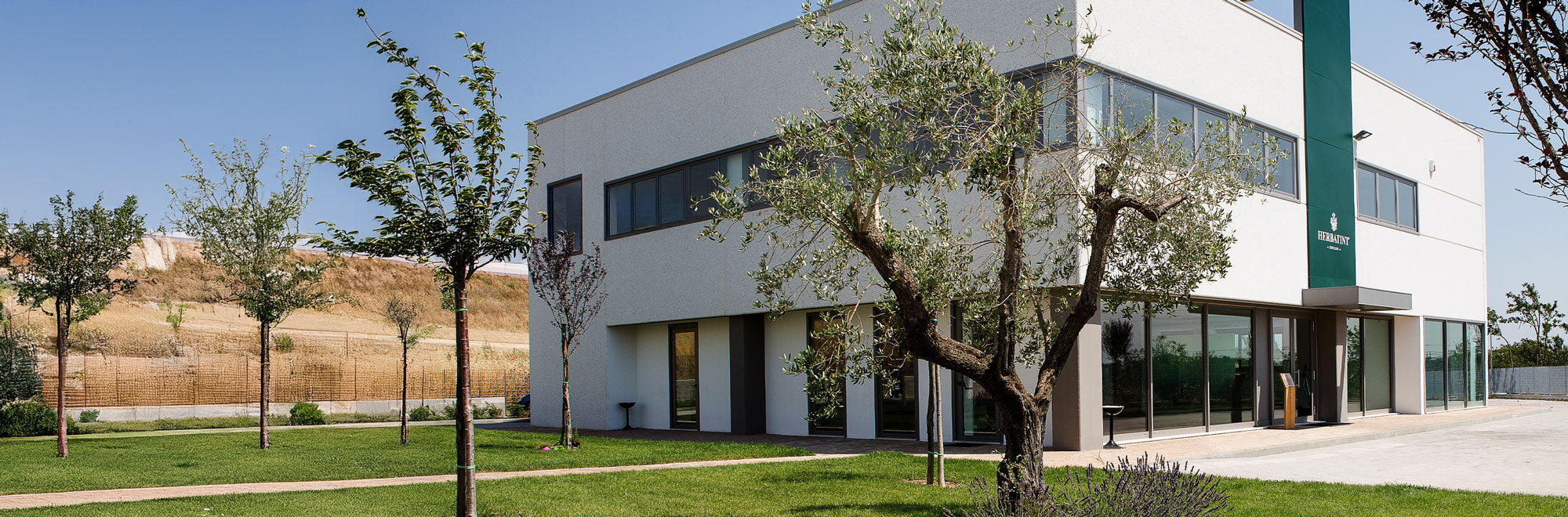 Veduta esterna dell’edificio principale Herbatint, circondato da alberi e spazi verdi ben curati, con il logo aziendale su una colonna verde verticale avanti l'ingresso