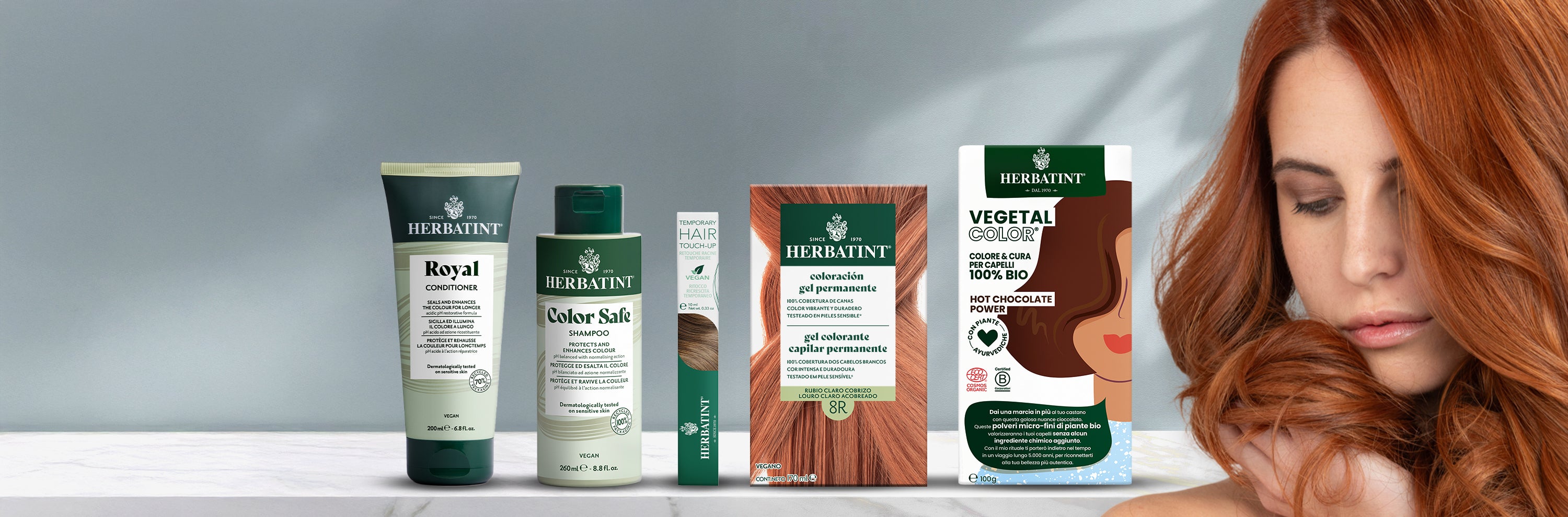 Línea completa de productos Herbatint sobre fondo neutro, incluidos champú, acondicionador, rímel para el cabello, geles colorantes permanentes y Vegetal Color. A la derecha, una mujer acaricia su largo cabello cobrizo.