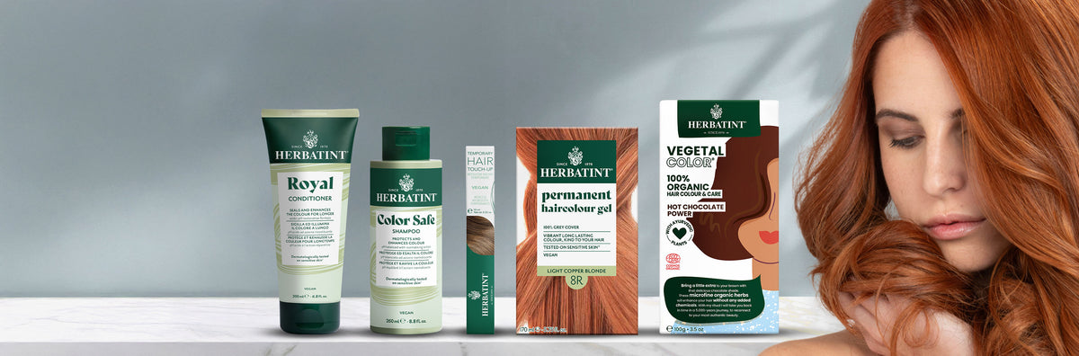 Linha completa de produtos Herbatint apresentada sobre fundo neutro, incluindo champô, amaciador, máscara para cabelo, géis de coloração permanente e Vegetal Color. À direita, uma mulher acaricia o seu longo cabelo ruivo.