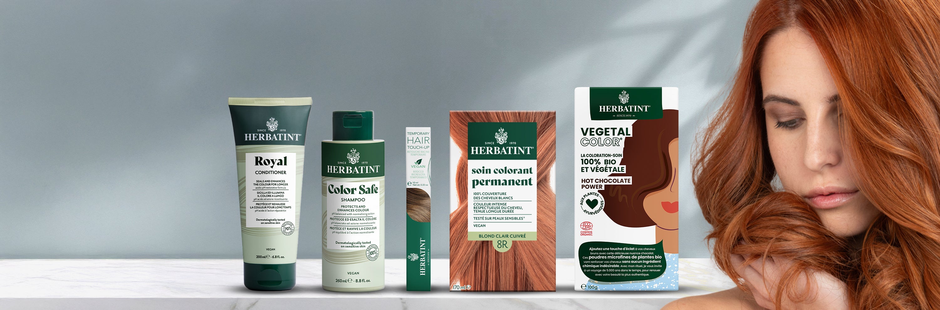 Gamme complète de produits Herbatint présentée sur fond neutre, incluant shampooing, après-shampooing, mascara pour cheveux, gels colorants permanents et Vegetal Color. À droite, une femme caresse ses longs cheveux cuivrés.