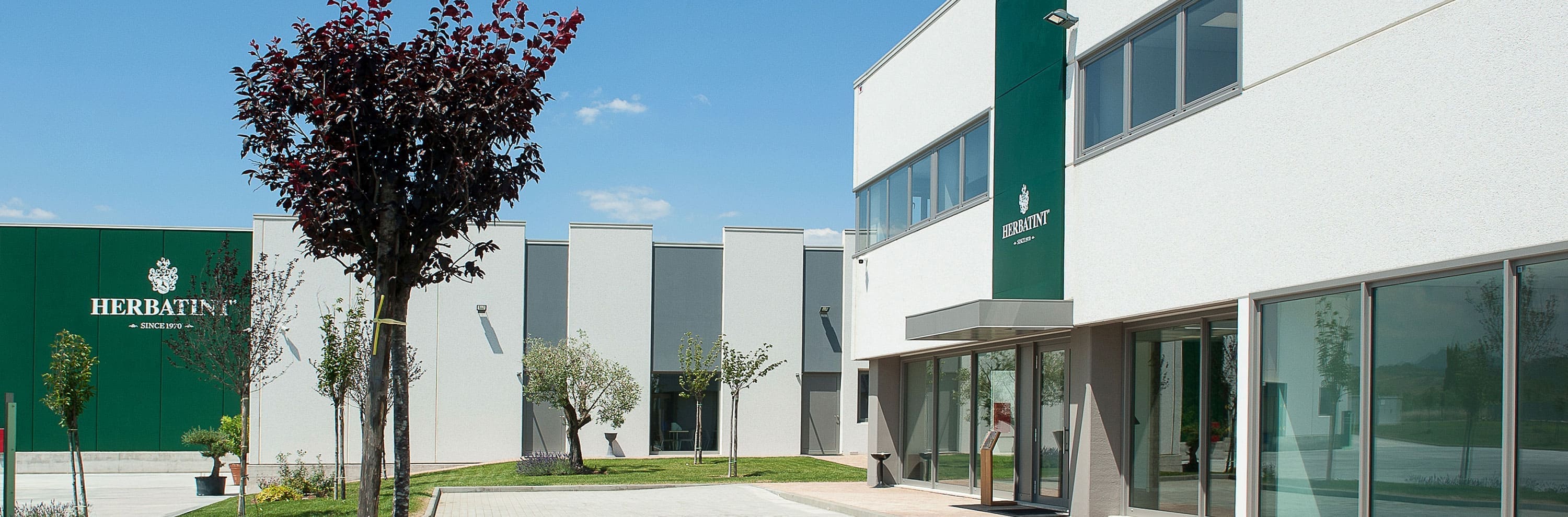 Panoramic view of the entrance to the Herbatint corporate buildings, featuring white and green facades, manicured green spaces, and decorative trees under a blue sky