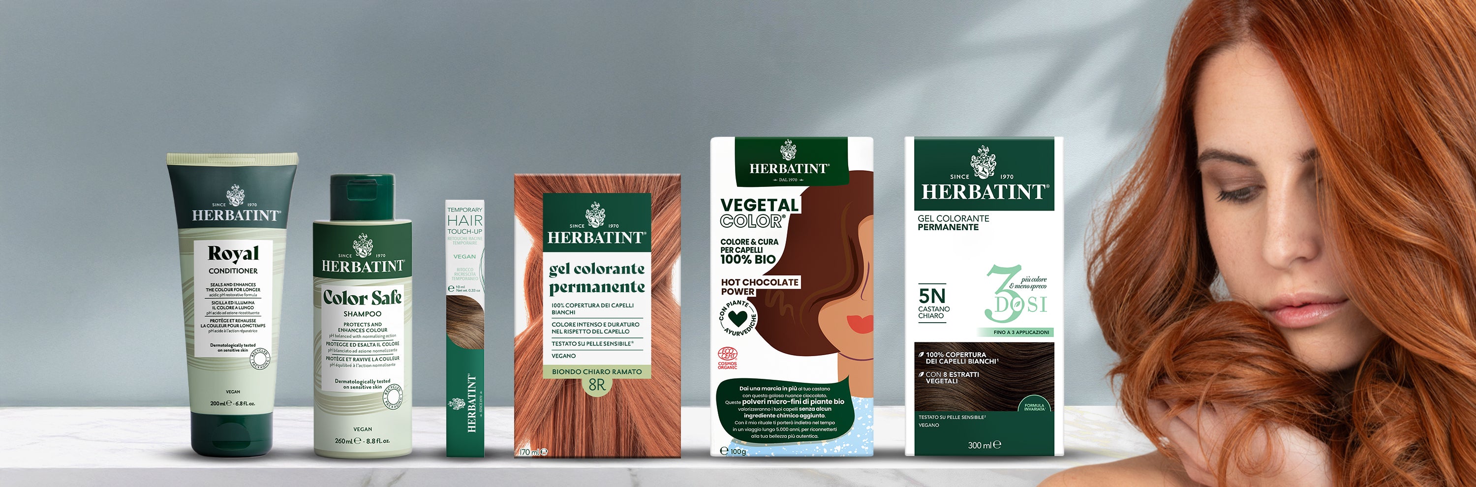 Linea completa di prodotti Herbatint esposta su fondo neutro, inclusi shampoo, balsamo, mascara per capelli, gel coloranti permanenti e vegetal color. A destra, una donna accarezza i suoi lunghi capelli ramati