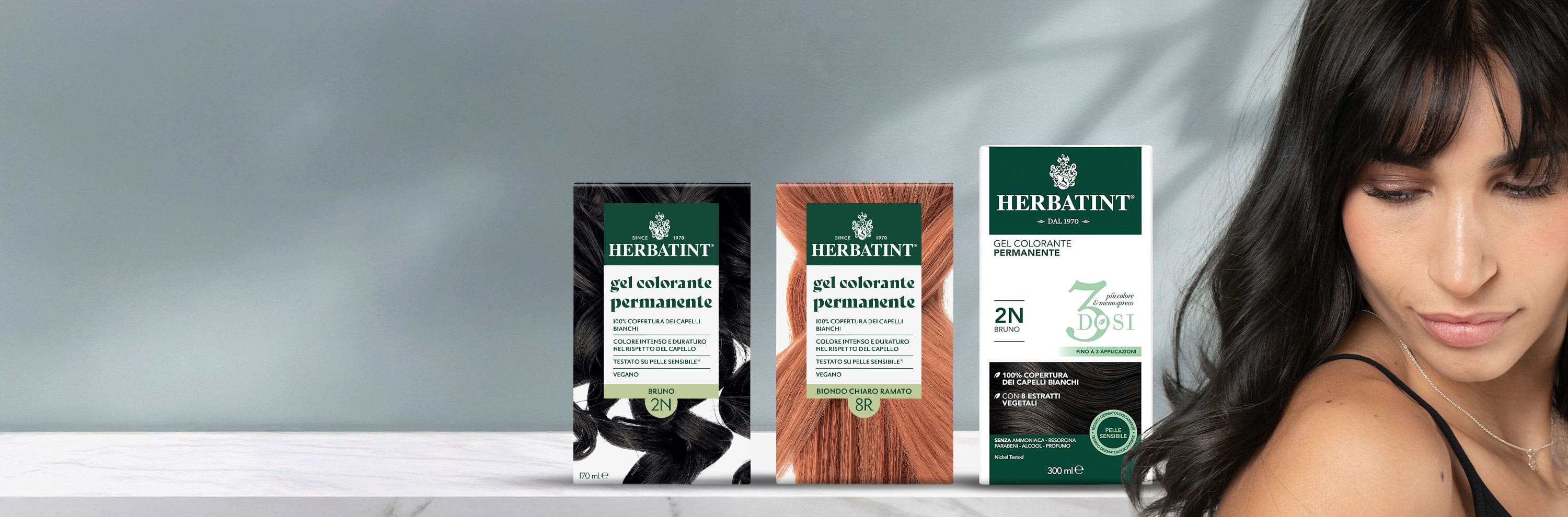 Tre confezioni di tinta Herbatint in diverse tonalità,bruno 2N e biondo chiaro ramato 8R, accanto a una donna con capelli scuri e lucenti