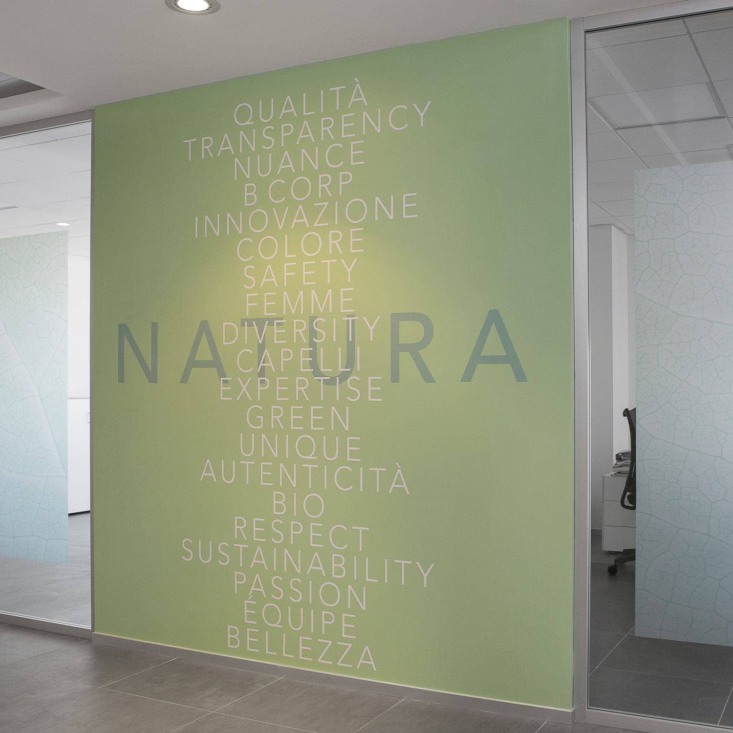 Parede verde decorativa no interior da sede da Herbatint com palavras-chave verticais em branco: “Sustainability”, “Passion”, “Beauty”, “Innovation” e “Expertise”, e “NATURE” a cinzento ao centro, representando os valores da empresa