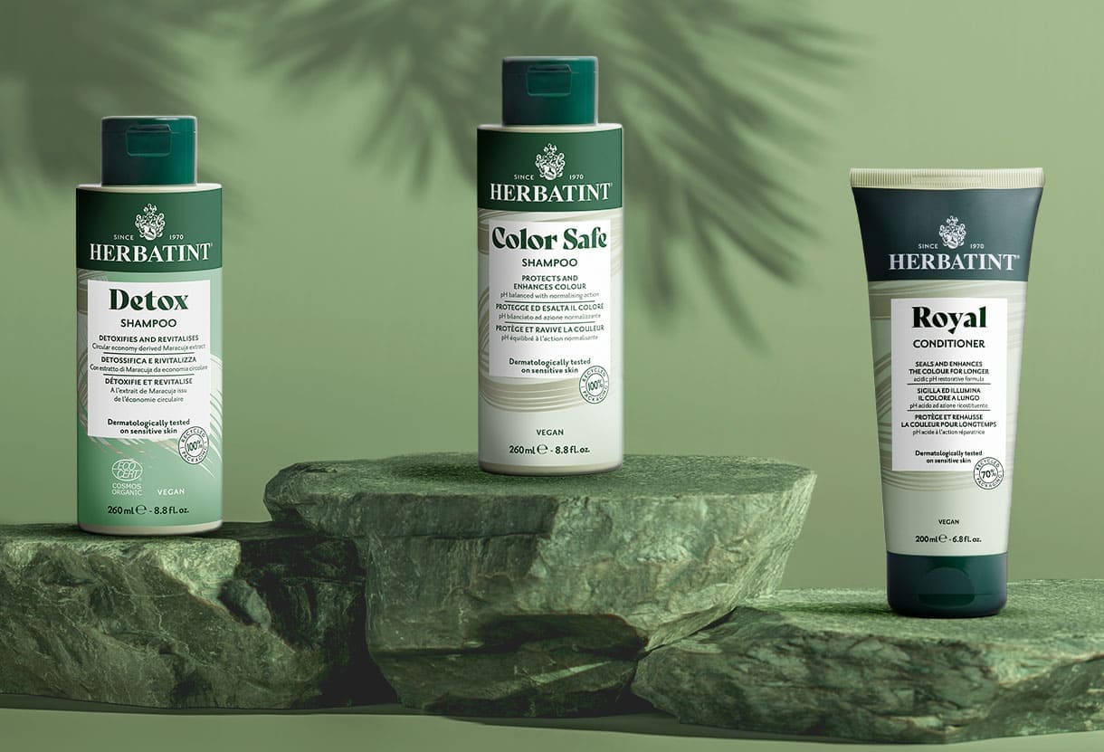 Produits de soin capillaire Herbatint — shampooing Detox, shampooing Color Safe, après-shampooing Royal — posés sur des pierres avec un fond vert naturel et une ombre de palmier